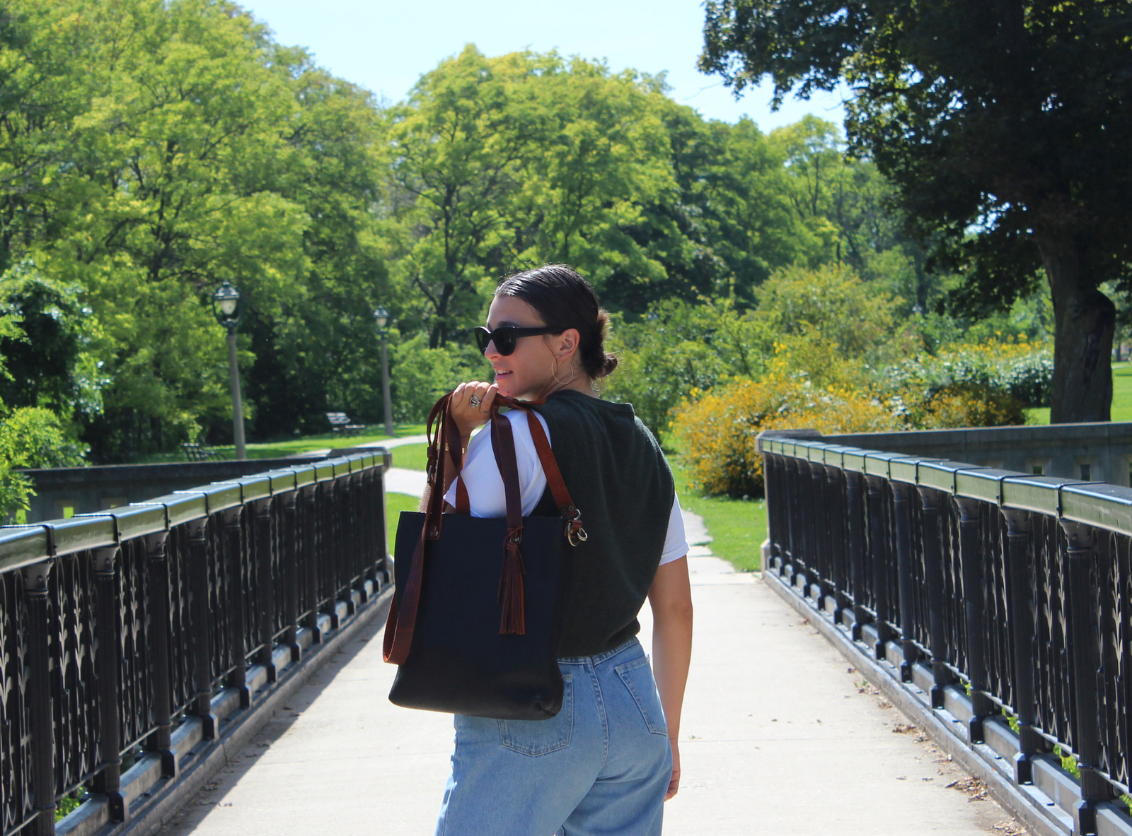 A woman stands in the center of a bridge and looks over her shoulder back at the camera. She wears blue jeans, a green sweater vest, and models a black and brown leather bag. 