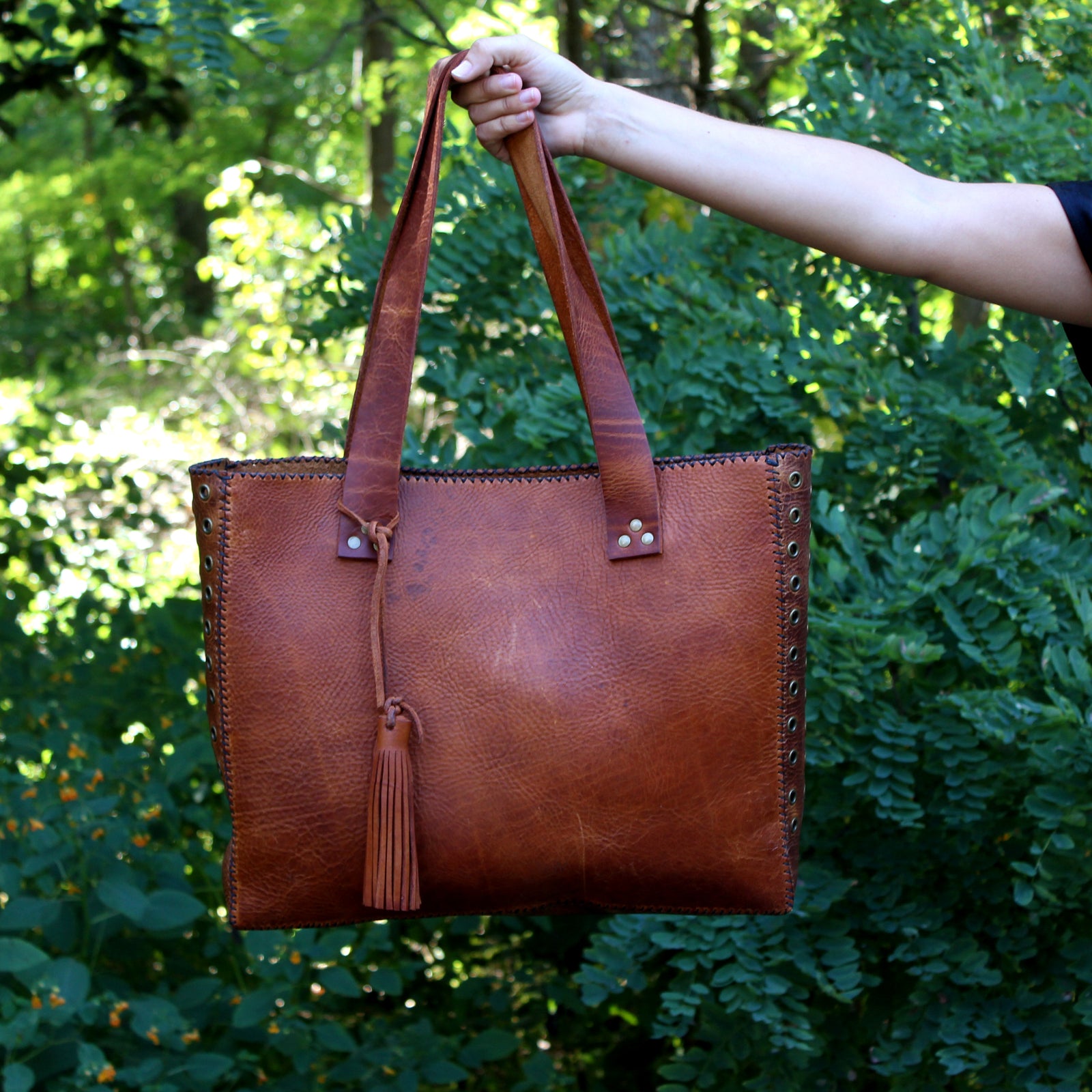 An outstretched arm holds a large tan leather bag in front of the camera.